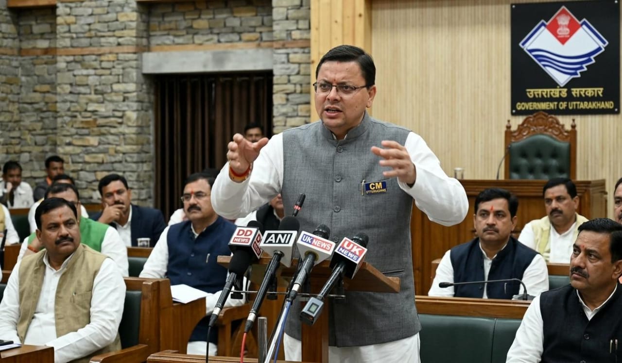 उत्तराखंड के मुख्यमंत्री पुष्कर सिंह धामी विधानसभा में पोडियम पर खड़े होकर दृढ़ता से भाषण देते हुए, पीछे सरकारी लोगो और अन्य विधायक बैठे हैं।